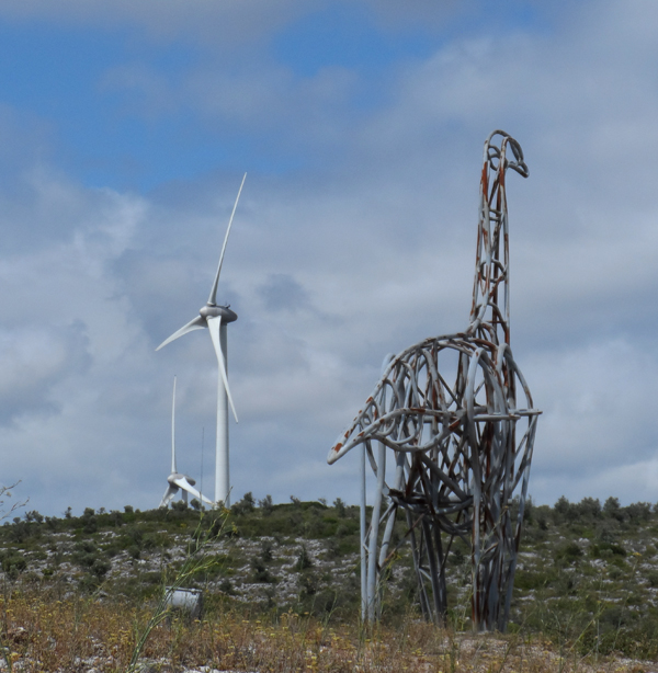 A metal dinosaur and wind turbine in a field set against a cloudy blue sky